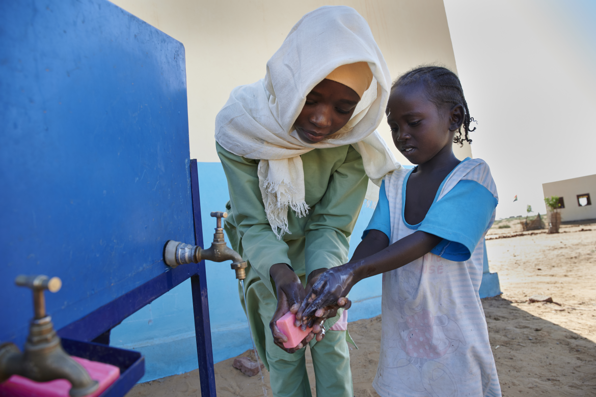 An older girl shows a kindergartner how to properly wash her hands using water and soap. Ban Jadid Primary School, El-Fasher, North Darfur, Sudan (2019). Credit: UNICEF/Noorani/UNI233850